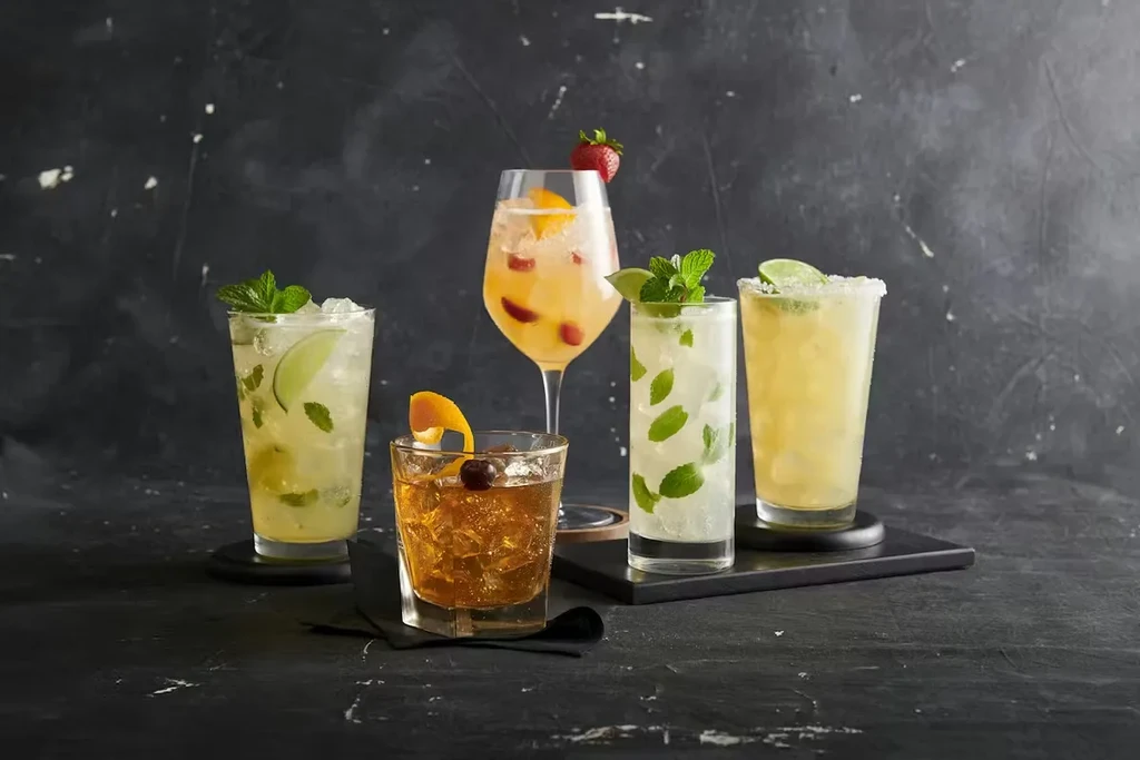 Five colorful cocktails with various garnishes against a dark background.