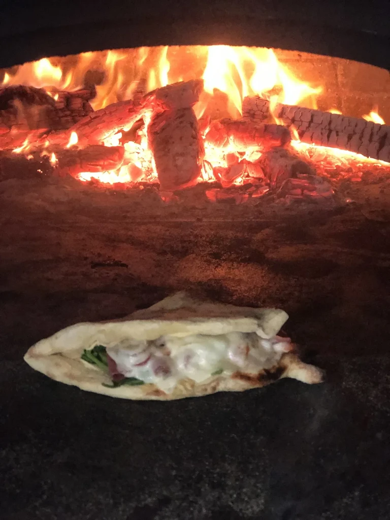 Wood-fired oven interior with vibrant flames cooking a filled flatbread.