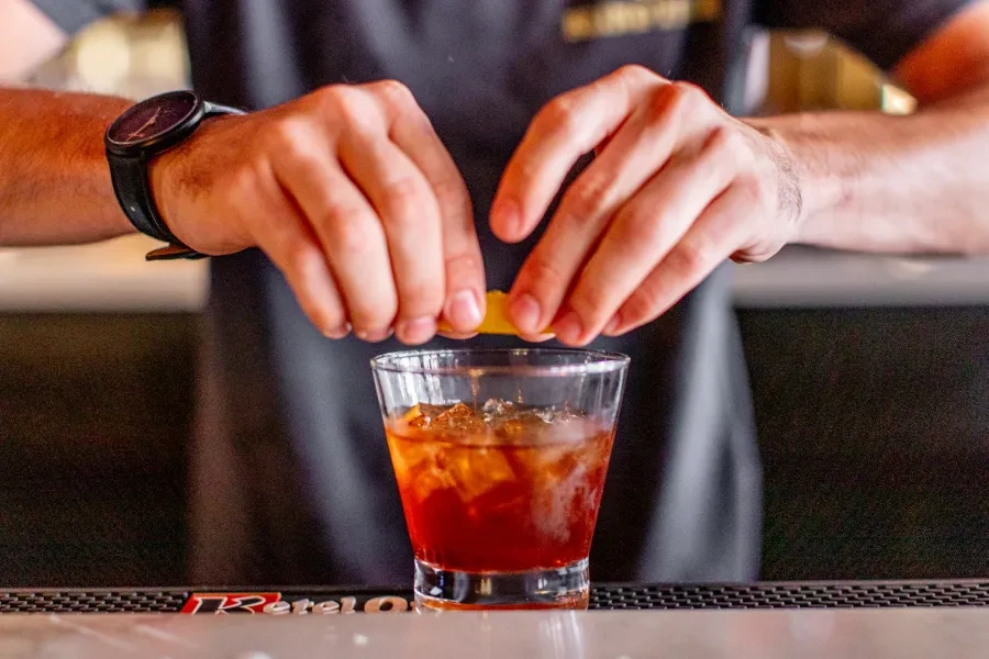 Hands squeeze orange peel over amber cocktail, creating a light mist.
