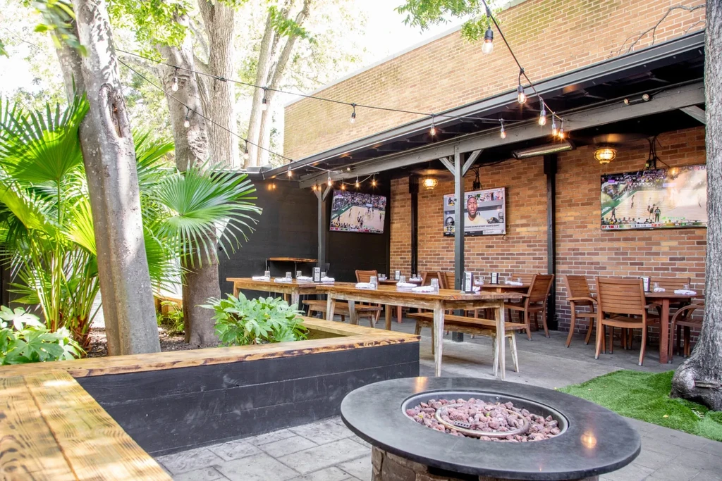 Outdoor restaurant patio with fire pit, dining tables, and TVs showing sports.