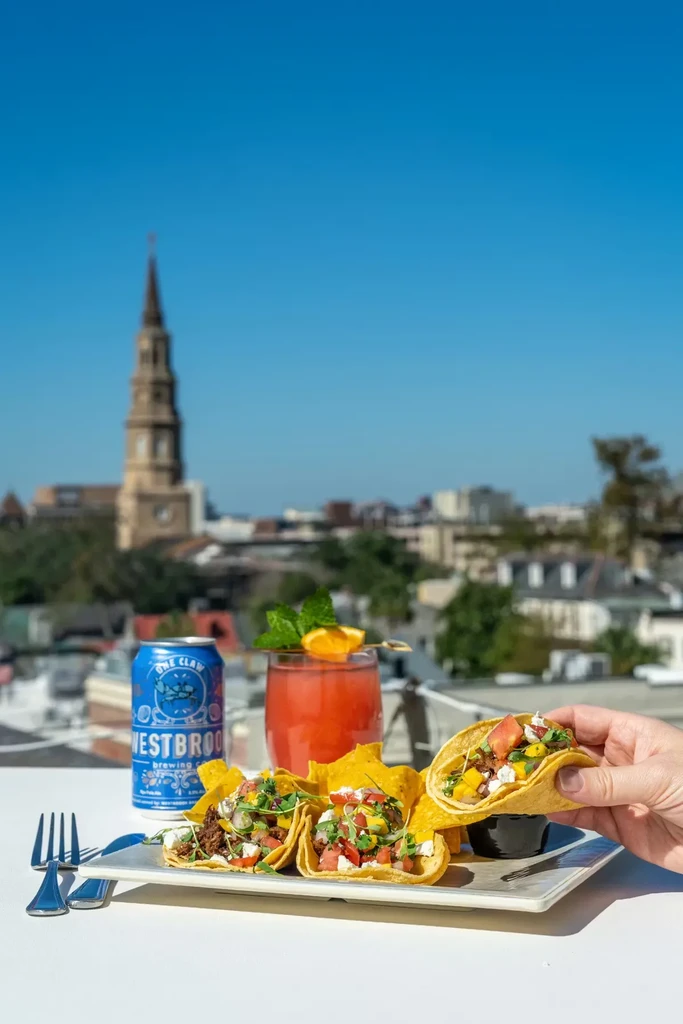 Hand holding a taco, cocktail, and beer can outdoors.