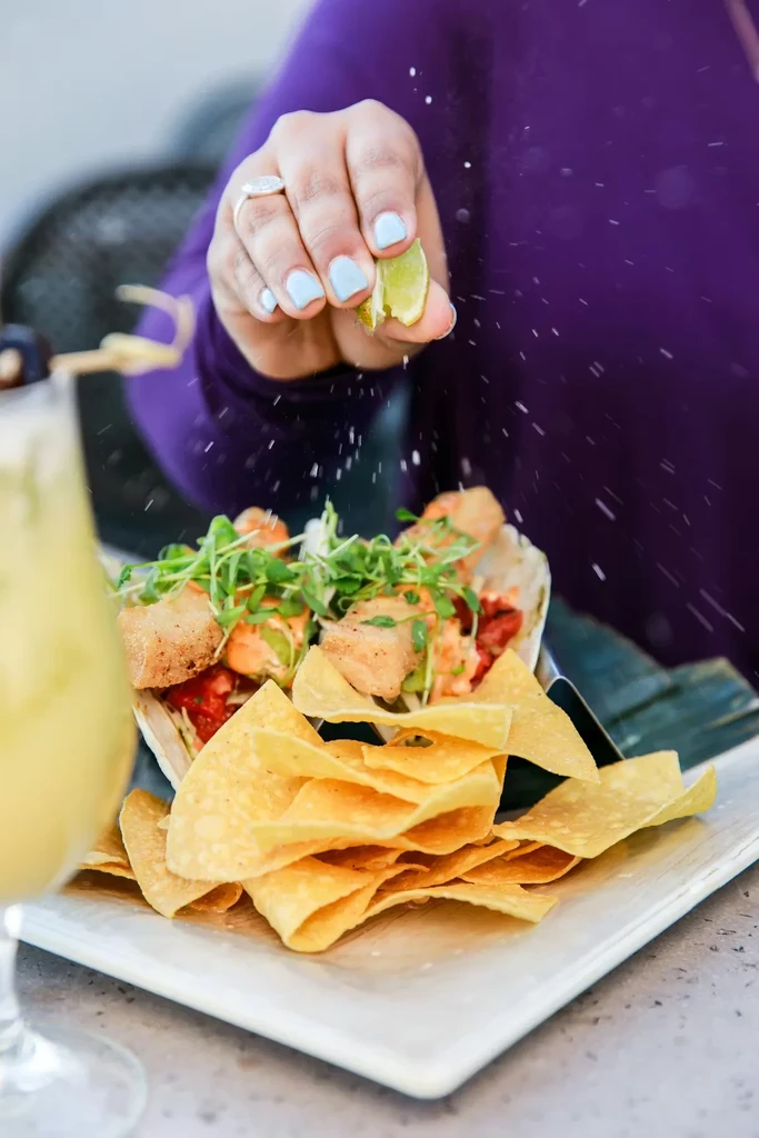 Hand squeezing lime over fish tacos with microgreens and chips.