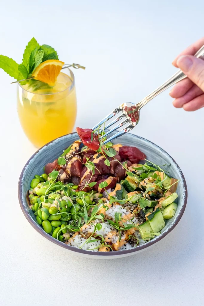 Hand with fork over a colorful tuna poke bowl and a citrus drink.