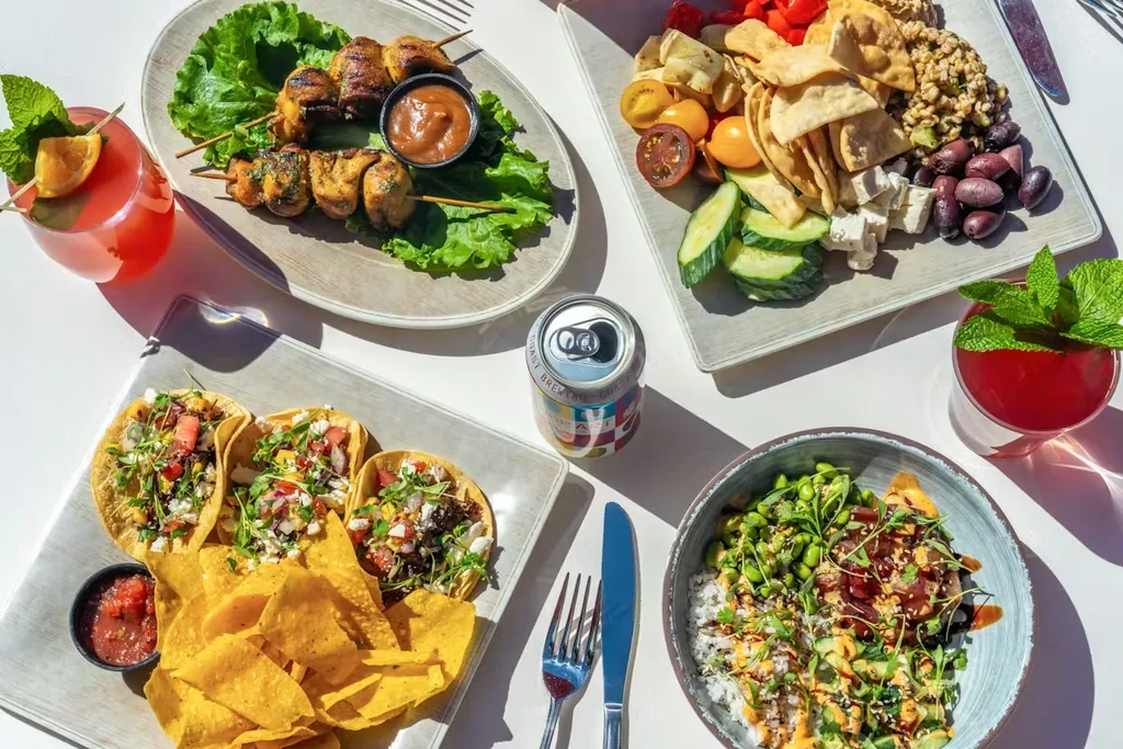 Flat-lay of skewers, tacos, salad, poke bowl, drinks on white table.