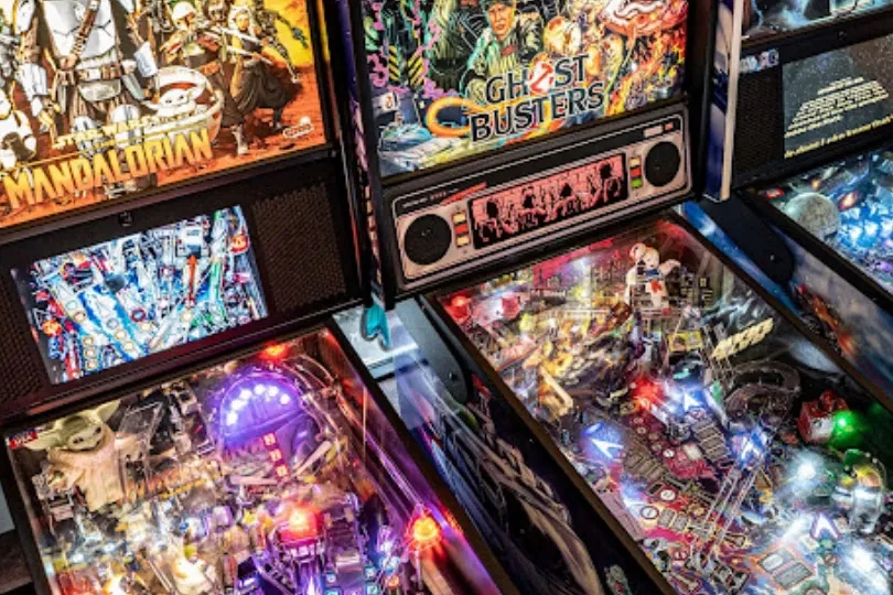Three brightly lit pinball machines: Mandalorian, Ghostbusters in an arcade.