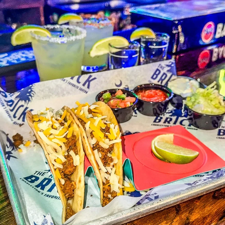 Hard-shell tacos, condiments, margaritas, and blue shots on a branded tray.