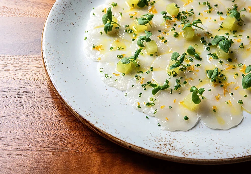 Thinly sliced white carpaccio with green herbs, zest on rustic plate.