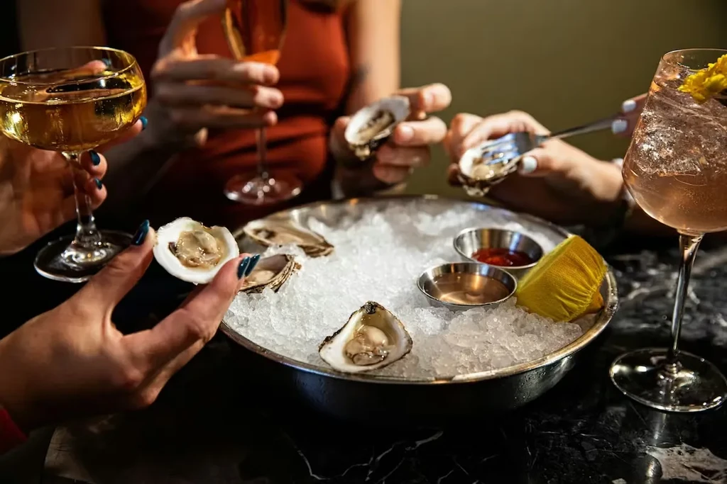 Multiple hands share a platter of raw oysters, sauces, and drinks.