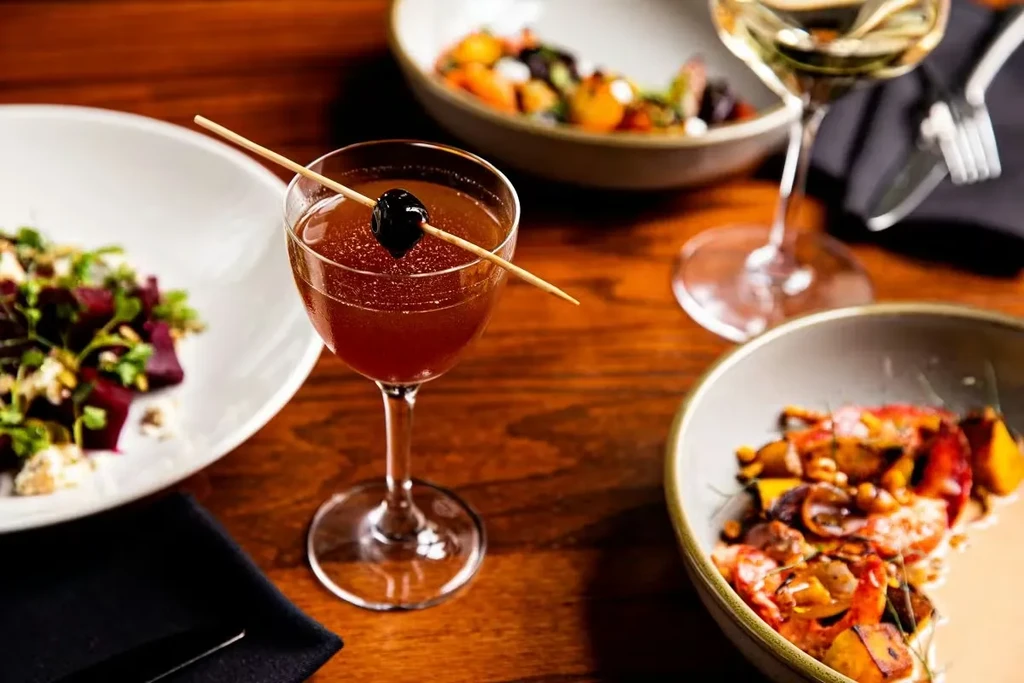 Close-up: Reddish-brown cocktail with cherry, surrounded by food on wood table.