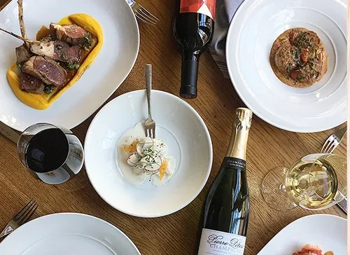 Flat lay: gourmet dishes, red wine, and champagne on a wood table.