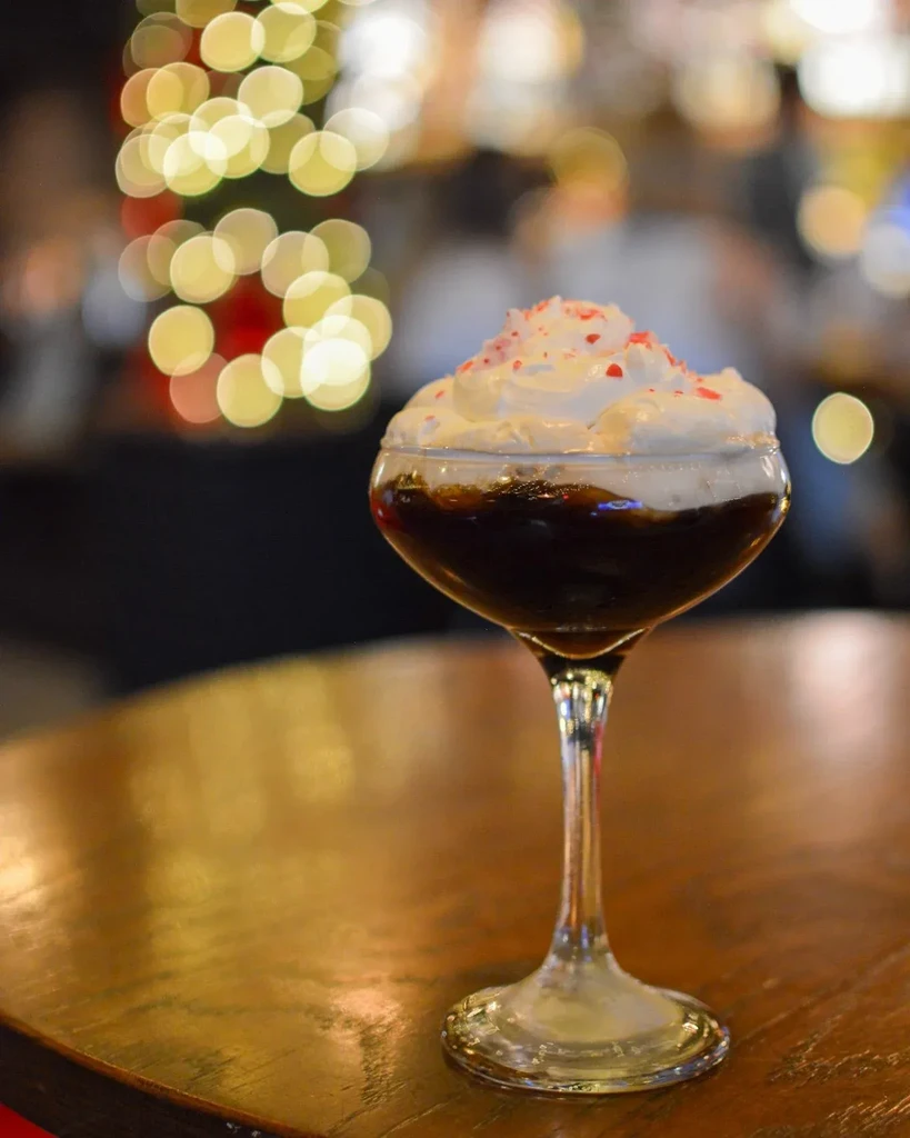 Festive dessert cocktail with whipped cream, red sprinkles, and bokeh lights.