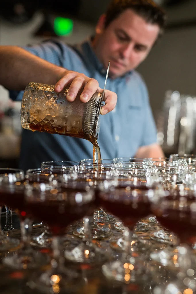 Hands pour a drink from a mixing glass into a line of coupes.