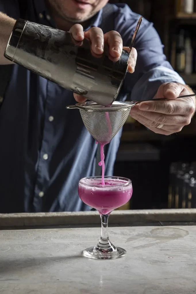 Bartender strains vibrant pink cocktail from shaker into a coupe glass.