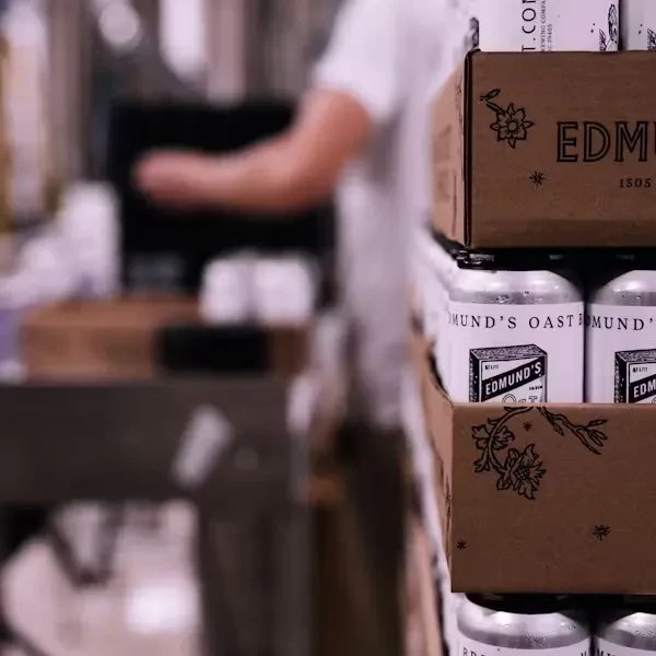 Close-up of stacked Edmund's Oast beer cans and boxes, person blurred.