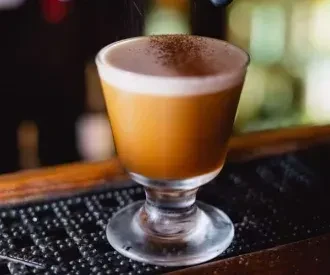 Amber cocktail in stemmed glass with frothy foam, dusted with spice.