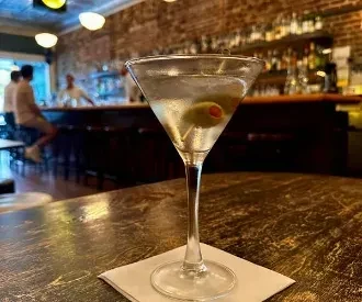 Condensing martini glass with olive on wooden table at blurred bar.