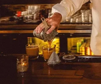 Bartender's hands pour an amber drink from a shaker into a glass.