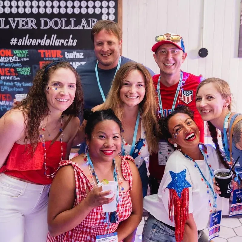 Seven smiling individuals in patriotic attire, holding drinks at a bar.