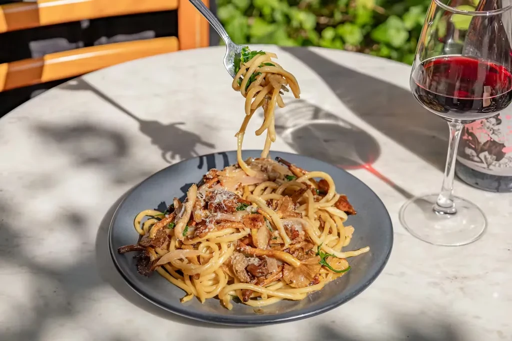Fork lifts pasta with mushrooms and herbs, red wine on marble.