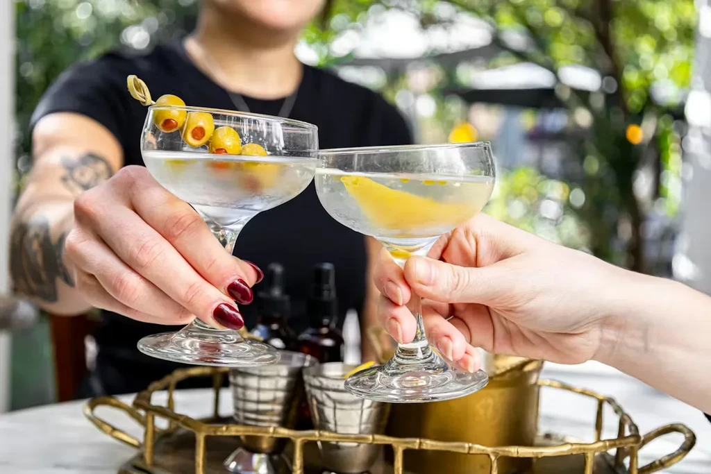 Two hands toast martinis with olives and lemon peel.