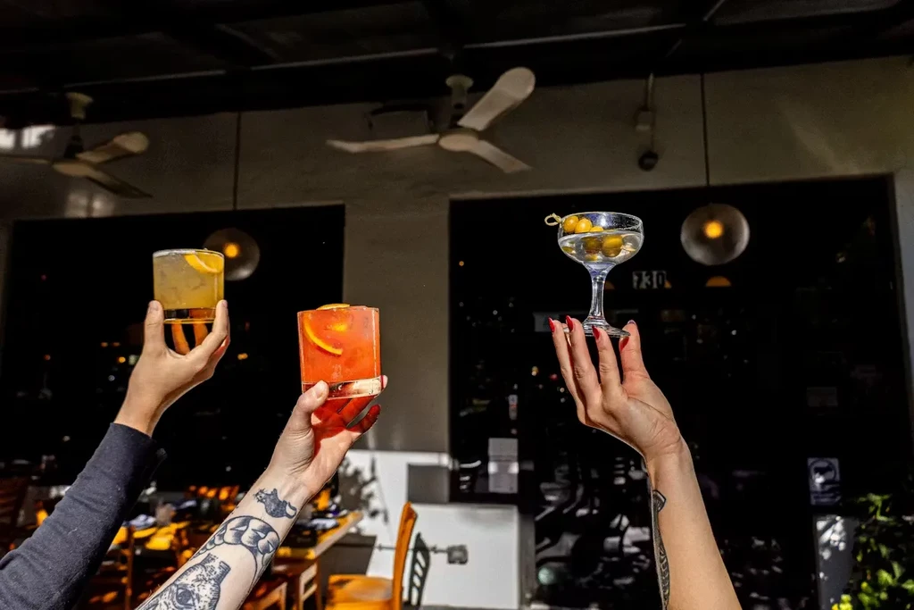 Three hands raise different garnished cocktails, toasting in a bar.