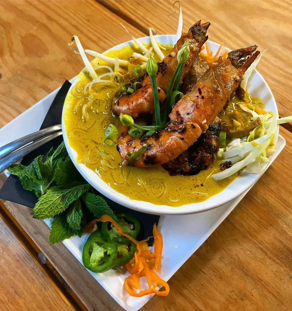 Vibrant prawn noodle curry dish with various garnishes on wood.