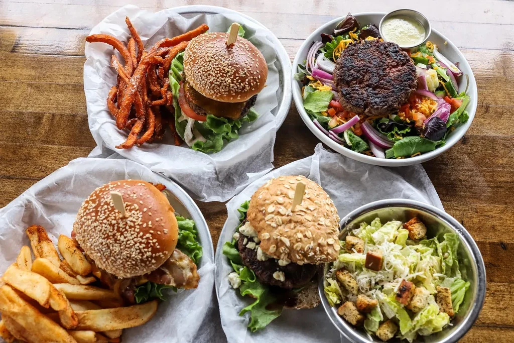 Top-down view of three burgers, two salads, and fries on a wooden table.