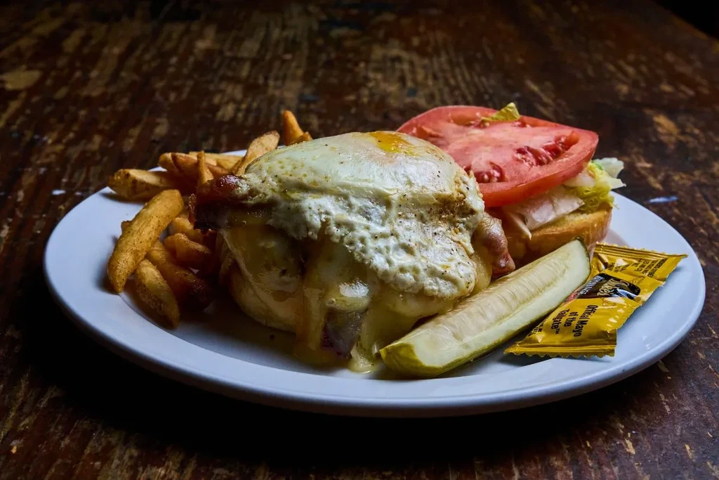 Burger, fried egg, melted cheese, fries, pickle on white plate.