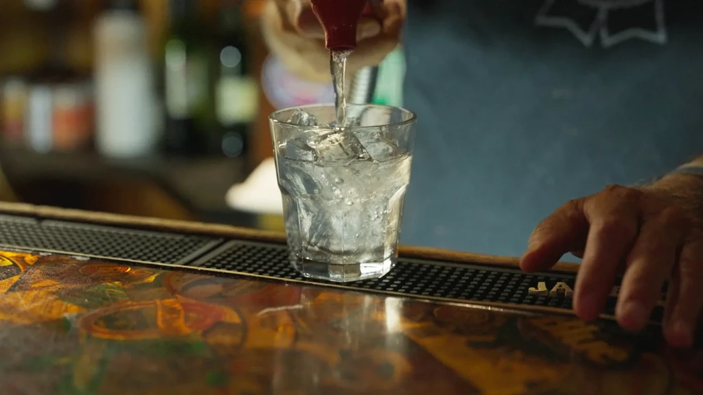 Hands pour clear liquid into an ice-filled glass at a bar.
