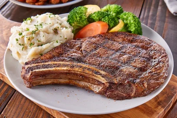 Seared bone-in steak, mashed potatoes, and colorful vegetables on plate.