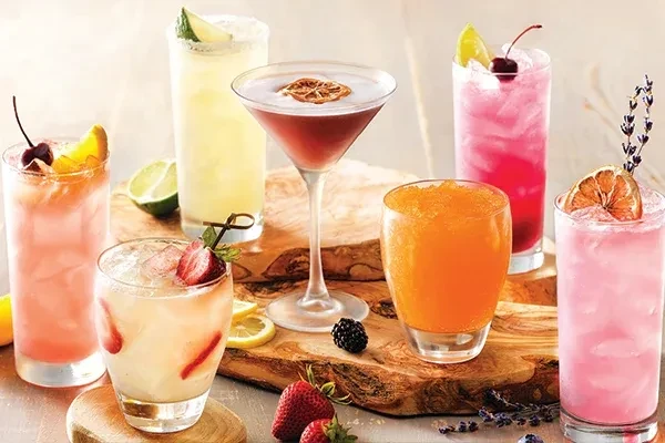Seven diverse, colorful cocktails with fruit garnishes on rustic wood.