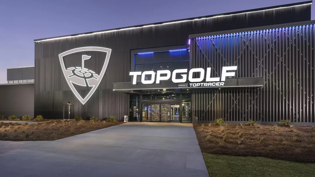 Modern Topgolf venue exterior at night with illuminated logos and blue lights.