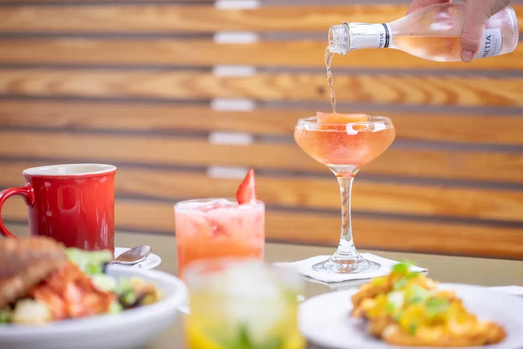 Hand pouring sparkling drink into glass with fruit; other drinks nearby.