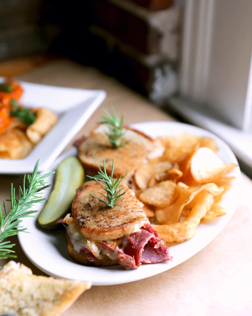 Reuben sandwich cut in half, served with chips, pickle, and rosemary.