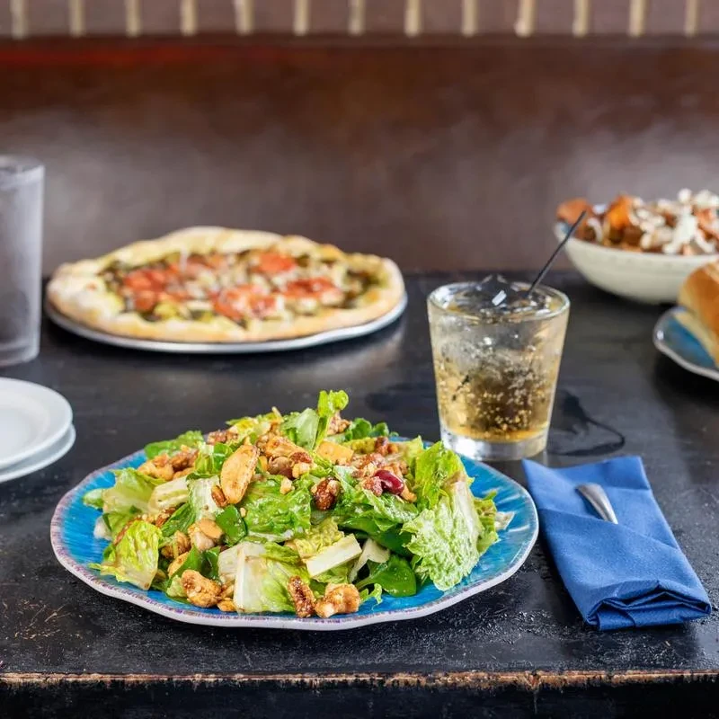 {A high-quality photo of restaurant dishes: salad, pizza, drinks on a dark table.}