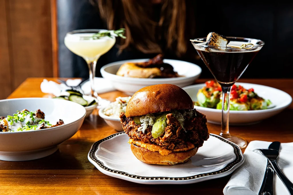 Crispy fried chicken sandwich, roasted vegetables, and cocktails on a restaurant table.