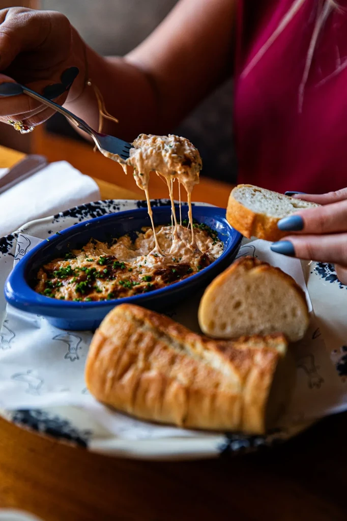 Hands pull stringy cheese from blue dip; bread poised for dipping.