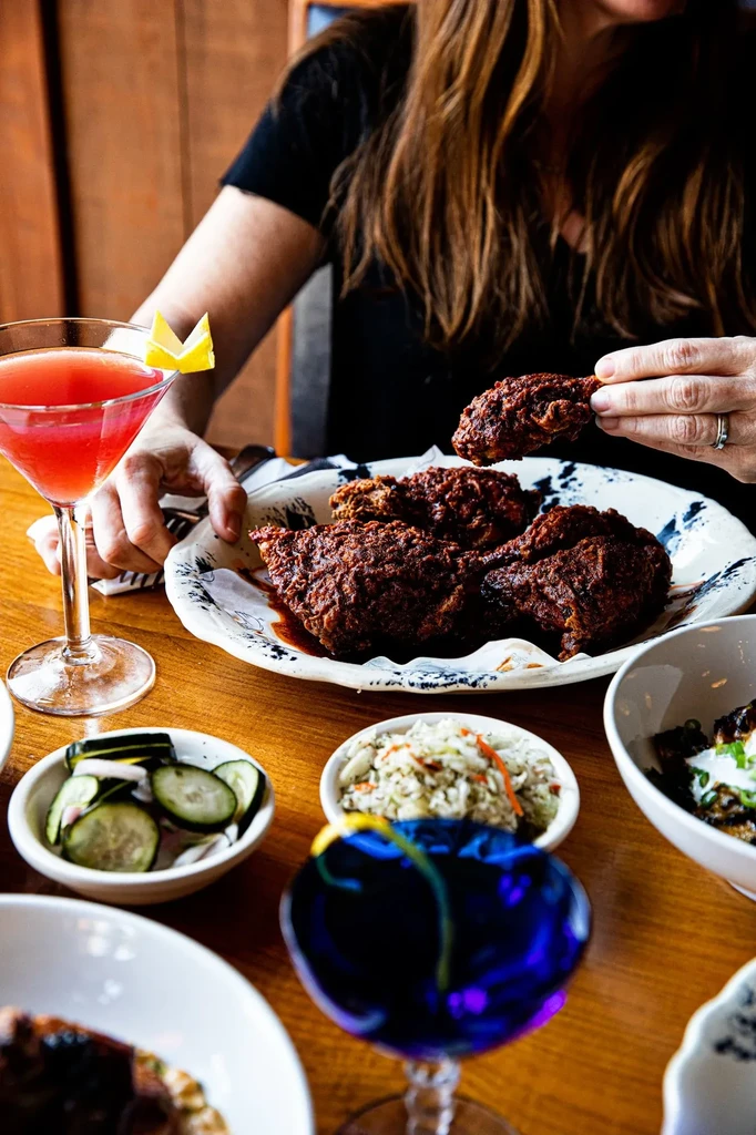 Hand lifting crispy fried chicken, sides, and two cocktails.