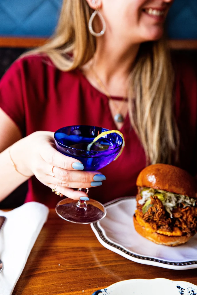 Blue cocktail and pulled-meat burger held by a woman's hand.