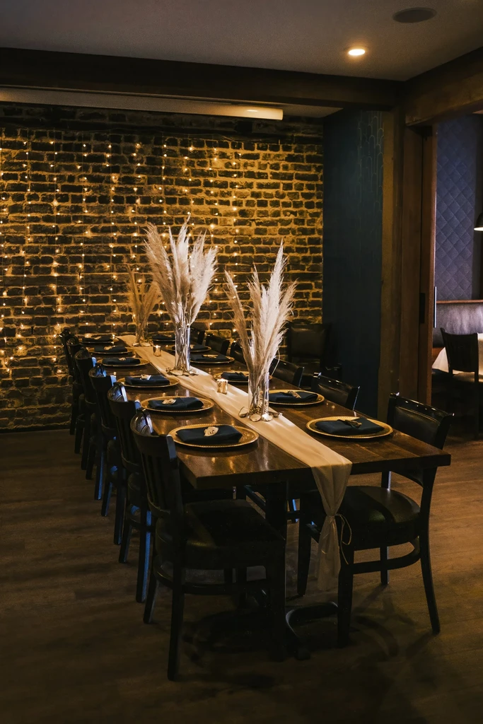 Dark wood dining table with gold chargers, pampas grass, and string lights.