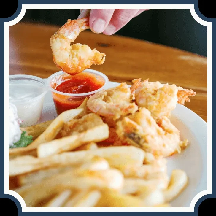 Hand dipping a fried shrimp into red sauce with fries.