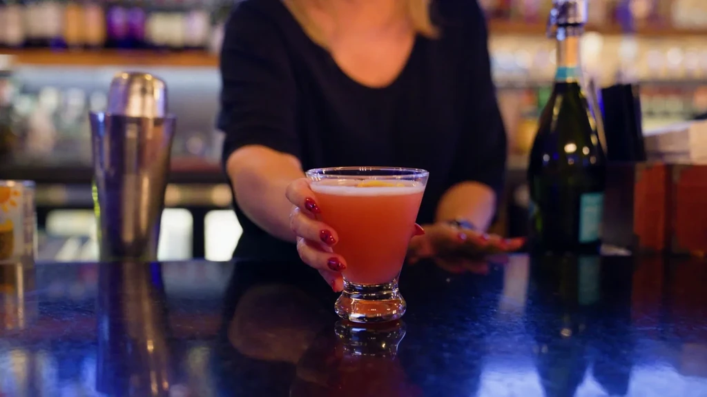 Hands hold a coral-pink cocktail with foam and citrus garnish in a bar.