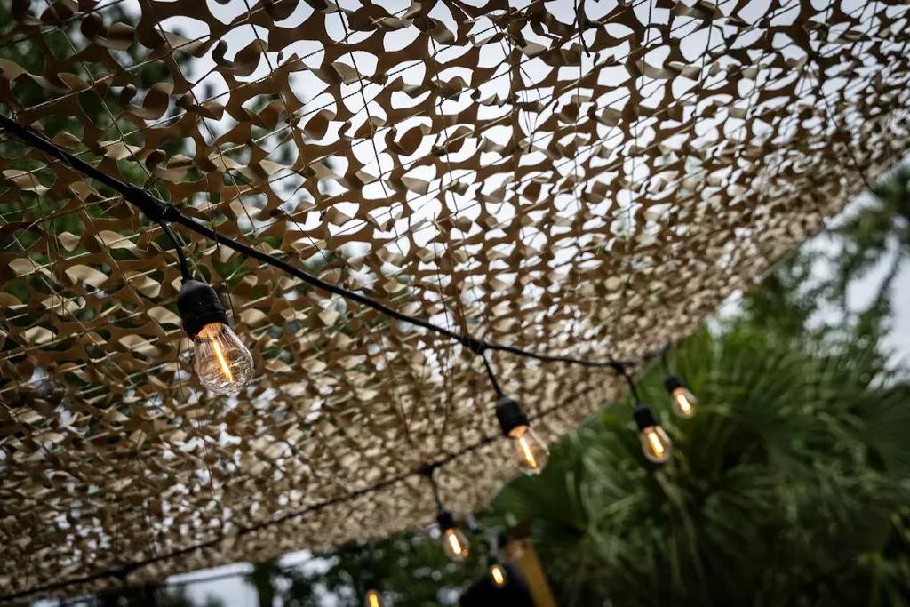 Glowing Edison bulbs under a camouflage net, with sky and foliage.