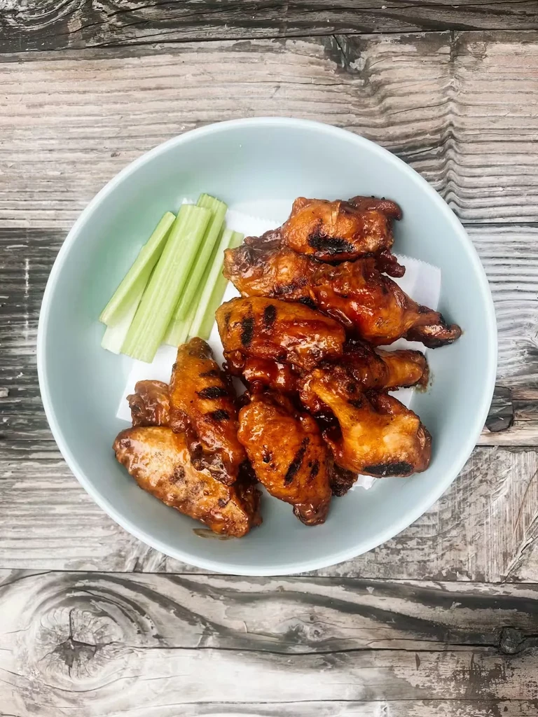 Bowl of saucy grilled chicken wings and crisp green celery on wood.
