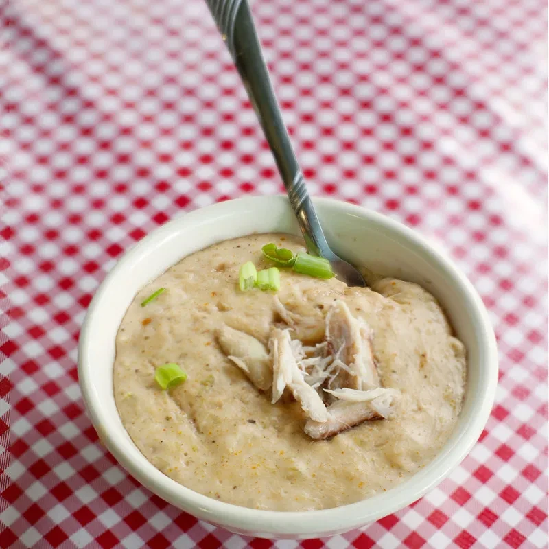 White bowl of creamy dish with shredded meat on checkered tablecloth.