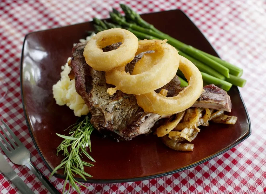 Grilled steak with onion rings, mashed potatoes, and asparagus on a checkered tablecloth.