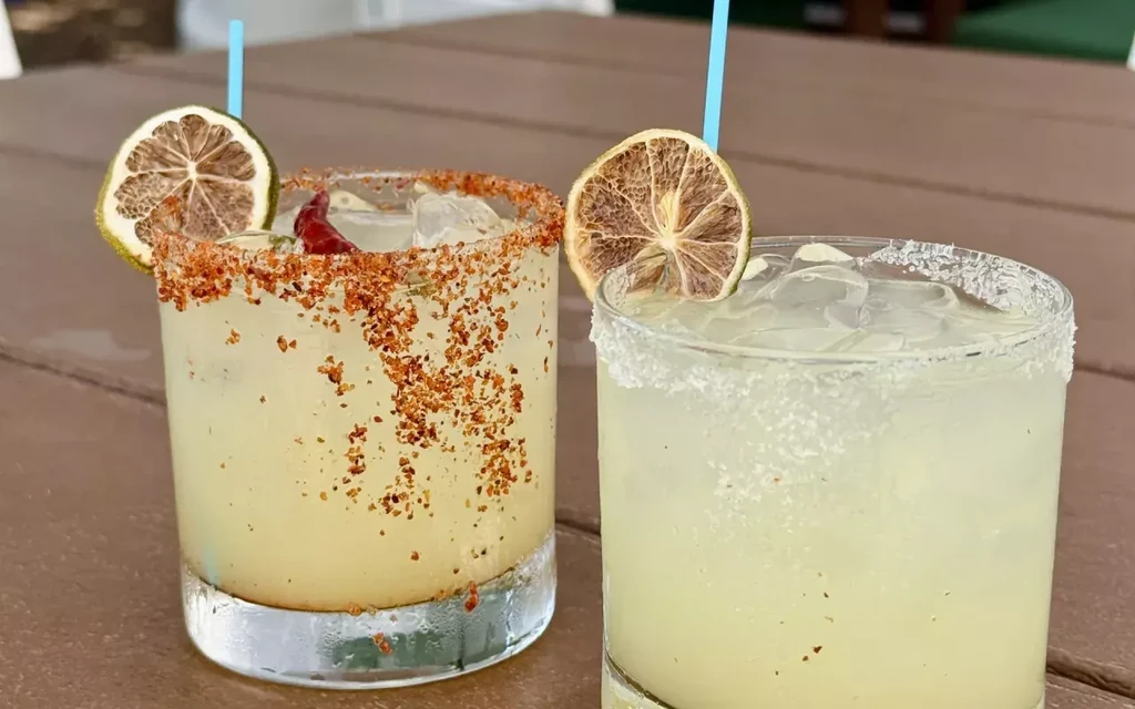 Two lime cocktails with ice, one chili-salted, garnished on wood.