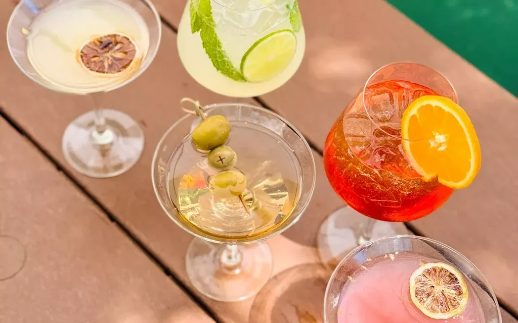 Overhead: Five garnished cocktails on a wooden deck near a pool.
