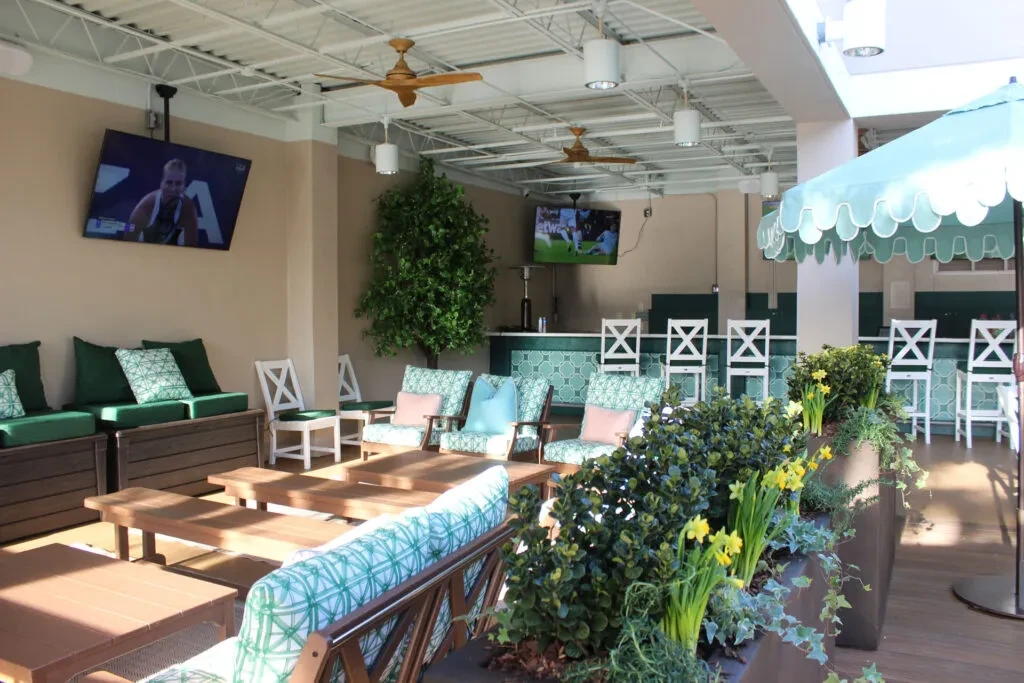 Modern restaurant patio with comfy seating, bar, TVs, and lush greenery.