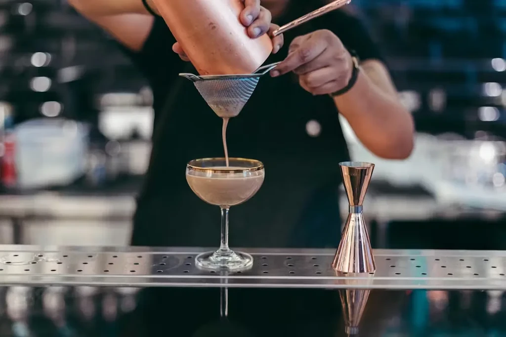 Hands pour creamy cocktail through strainer into a stemmed glass.